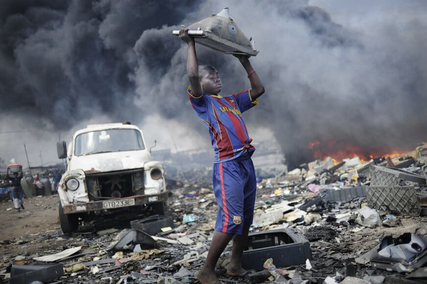 © Kai Löffelbein/laif, Afrika, Ghana, Accra, Agbogbloshie, 20.09.2011. Ein Junge wirft einen Fernseher mehrere Male auf den Boden, um an das Metall zu kommen. Im Hintergrund werden Reifen verbrannt.